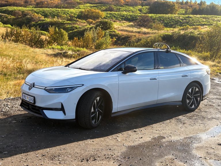 Hvítur Volkswagen ID.7 leigubíll í haustbirtu með grænni brekku í bakgrunni.
