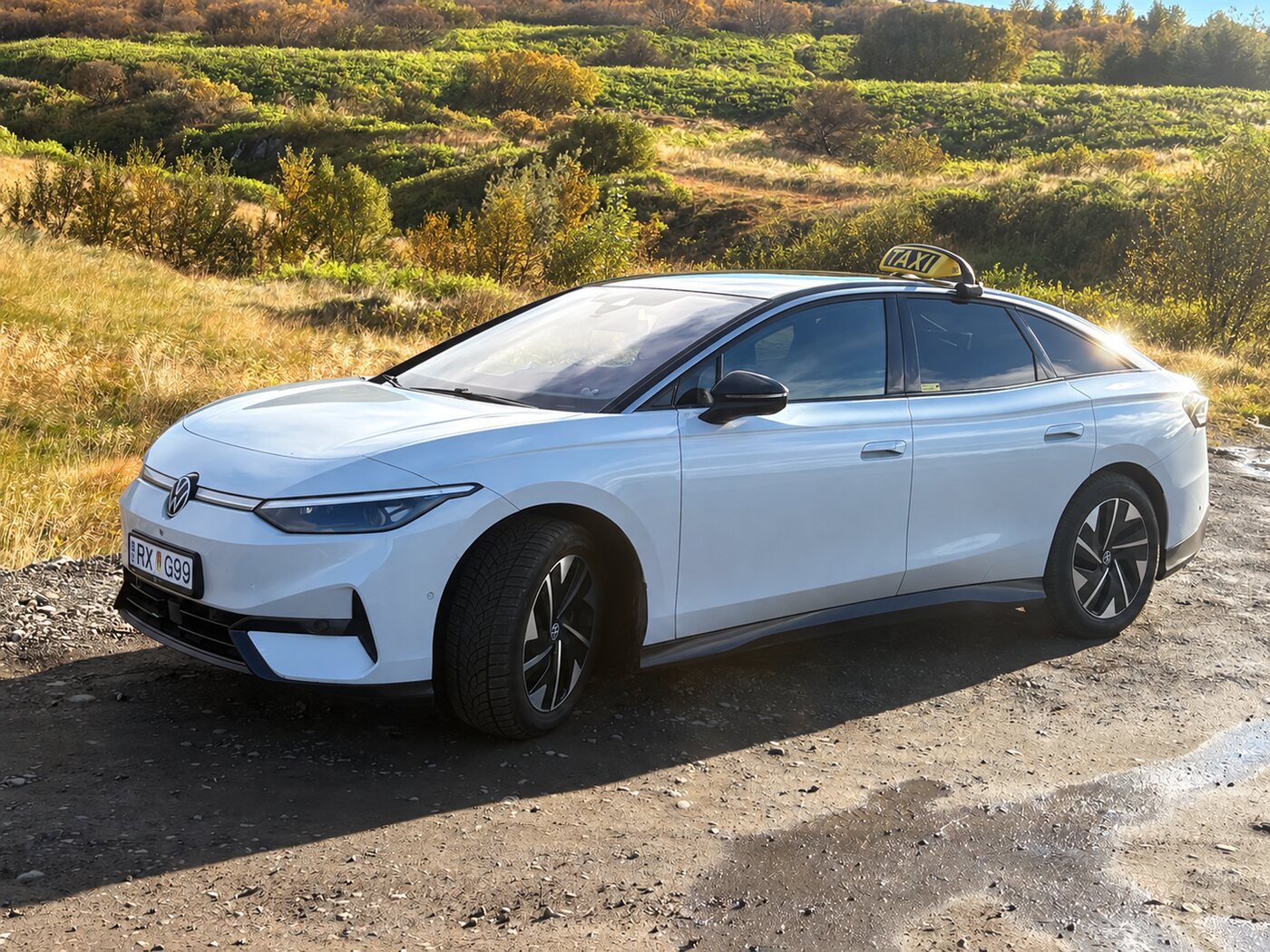 Hvítur Volkswagen ID.7 leigubíll í haustbirtu með grænni brekku í bakgrunni.